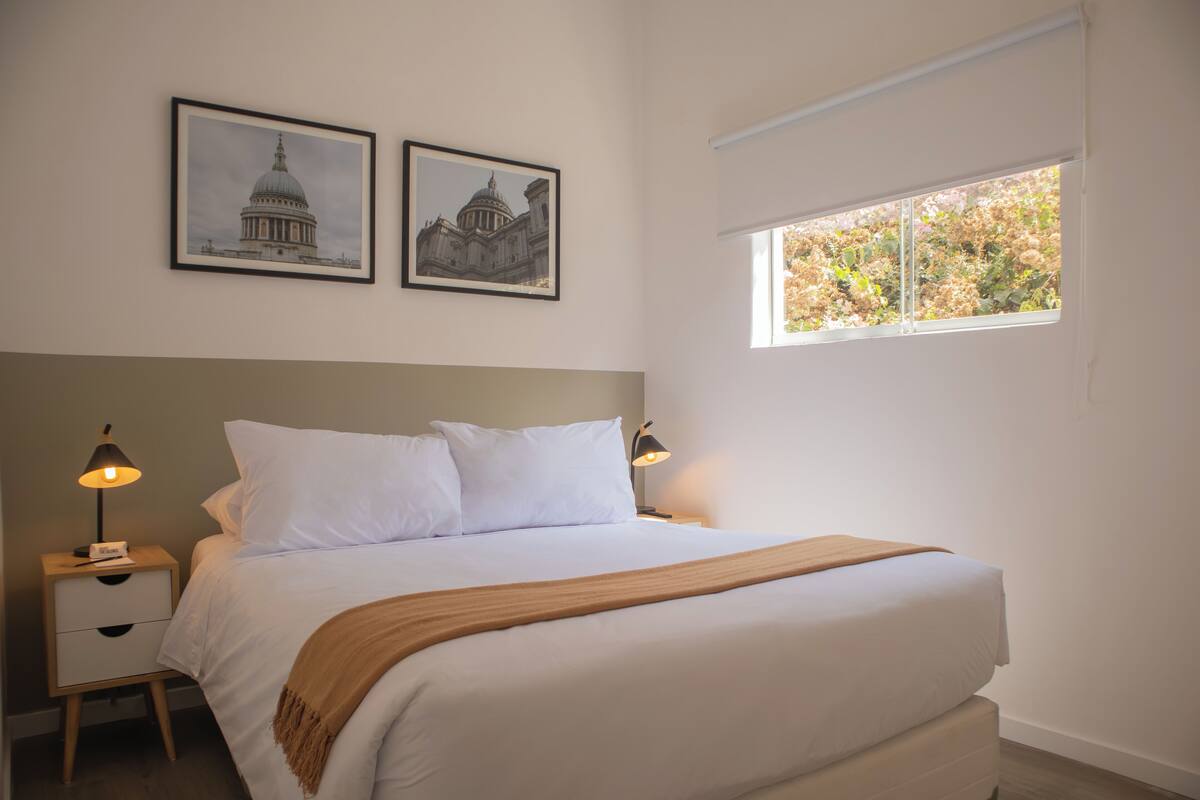 Un dormitorio con una cama grande, dos mesitas de noche con lámparas, una ventana que da a vistas de árboles y dos cuadros en la pared.