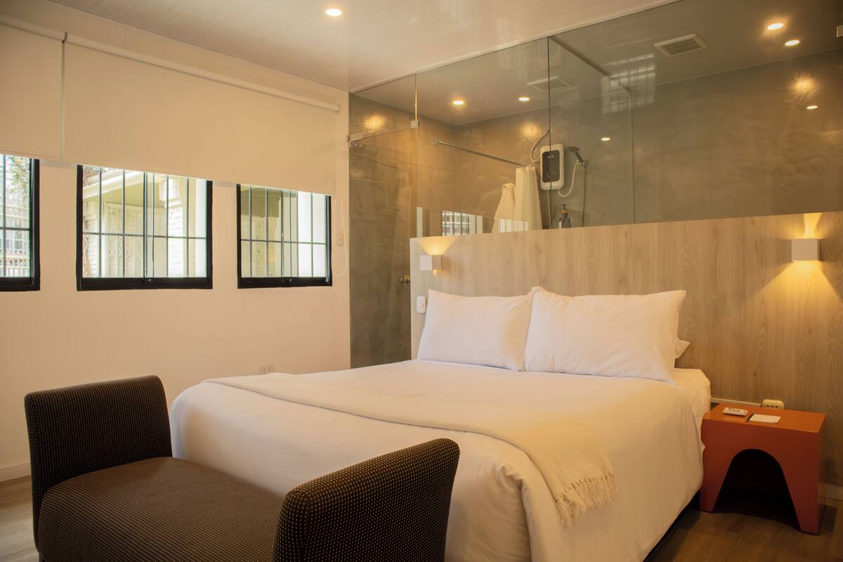 Una habitación de hotel moderna con una cama grande, dos sillones, cabina de ducha de vidrio y cabecero de madera.