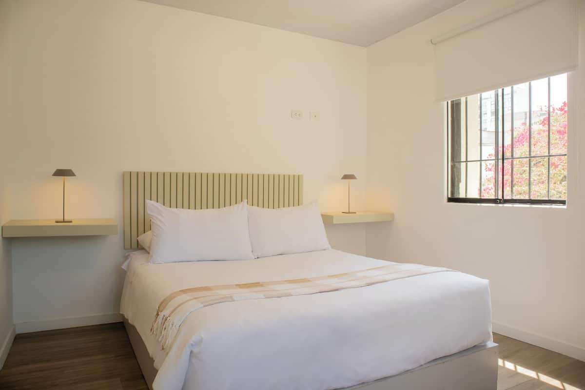 Un dormitorio ordenado con cama, dos mesitas de noche y una ventana.