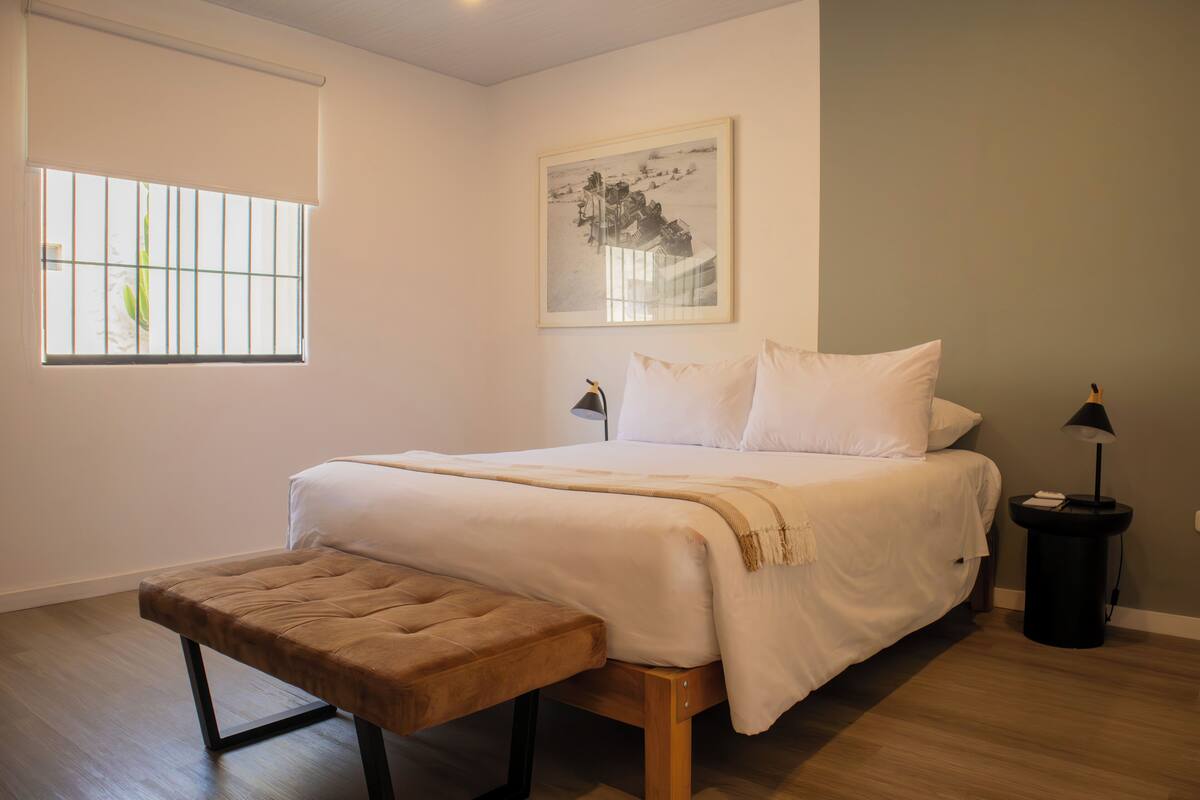 Un dormitorio con una cama, un banco, una ventana, un cuadro enmarcado y una mesita de noche.