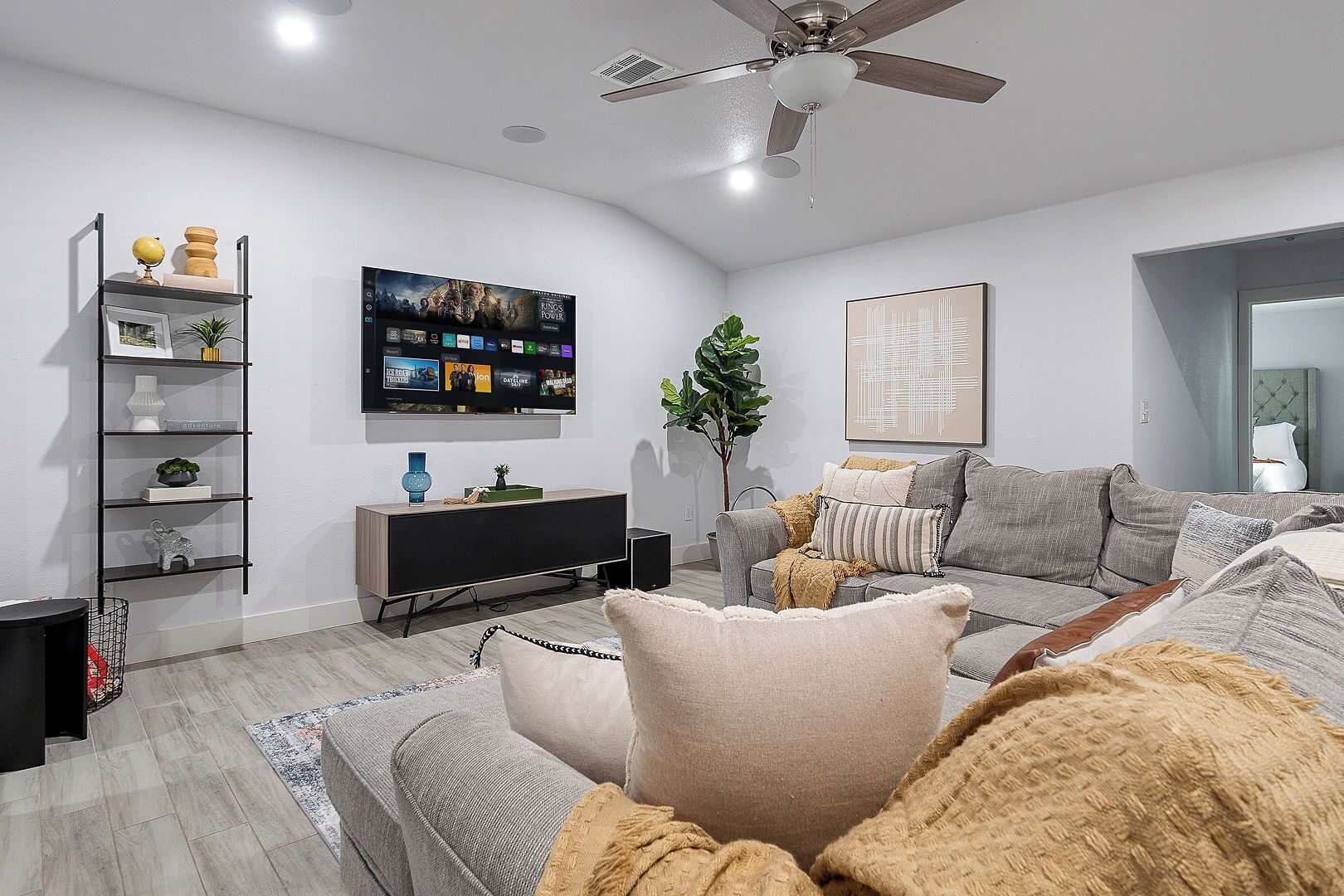 A modern living room with a grey sofa, a black entertainment unit, a ceiling fan, and a large flat-screen TV mounted on the wall.