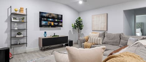 Un salon moderne avec un canapé gris, un meuble de rangement noir pour appareils électroniques, un ventilateur de plafond et un grand téléviseur à écran plat fixé au mur.
