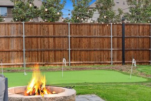 A backyard with a fire pit, artificial grass, a wooden fence, and string lights.