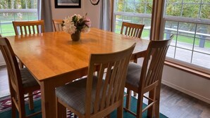 A dining room with a wooden table and six chairs, a vase with flowers, and large windows with a view of greenery.