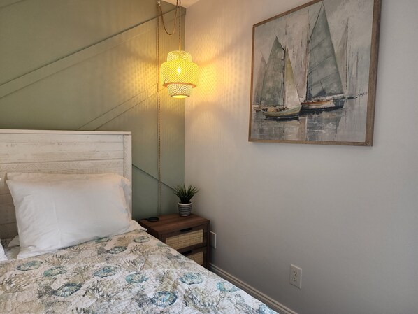 A bedroom with a bed, a nightstand, a wall-mounted lamp, a small potted plant, and a painting of sailboats. - Dogs Welcome-Close to Beach-Fenced Yard (Sebastian)