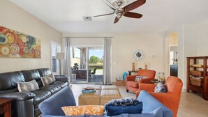 A spacious living room with a black leather sofa, an orange armchair, and a blue sofa. There is a ceiling fan, a large abstract painting, and a clock on the wall. - Cozy 3 bedroom home with private heated pool on a golf course with mountain view (Avondale)