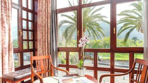 A room with large windows, a bench, two chairs, a small table with a plant, and a view of palm trees and greenery. - Indiana Boutique Hotel (Llanes)
