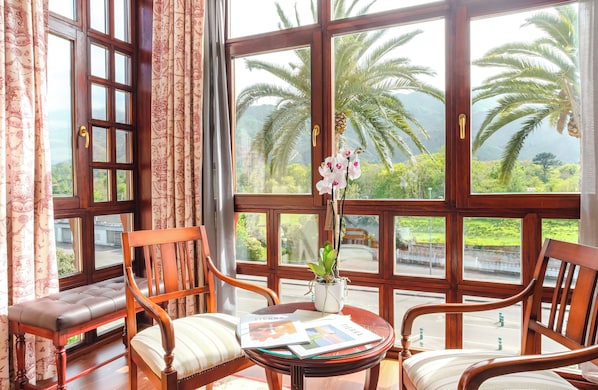 A room with large windows, a bench, two chairs, a small table with a plant, and a view of palm trees and greenery. - Indiana Boutique Hotel (Llanes)