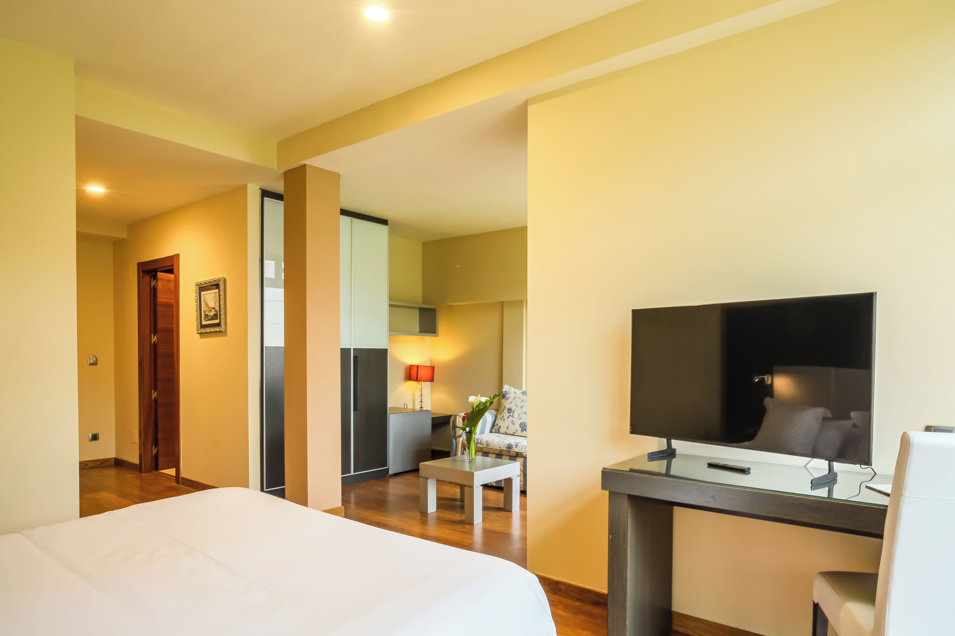 A hotel room with a bed, a desk with a chair, a TV, and a small table with a lamp and flowers.