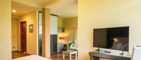 A hotel room with a bed, a desk with a chair, a TV, and a small table with a lamp and flowers.