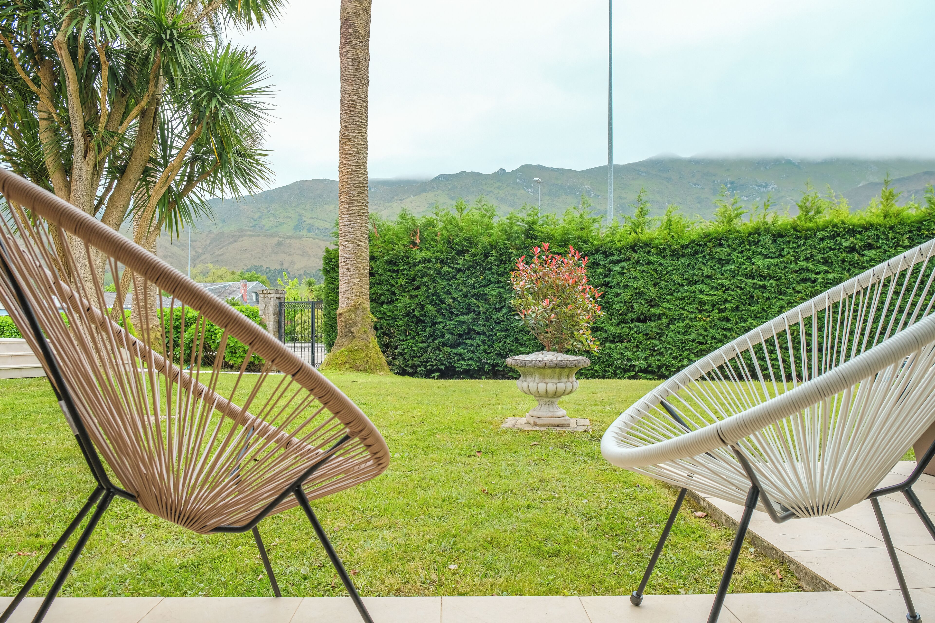 Dos sillas de mimbre mirando un jardín con una fuente, palmeras y vistas a colinas distantes.