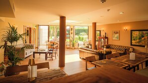 A spacious hotel lobby with a sofa, dining tables, and chairs, large windows with a view of greenery, and a mix of modern and classic decor. - Indiana Boutique Hotel (Llanes)
