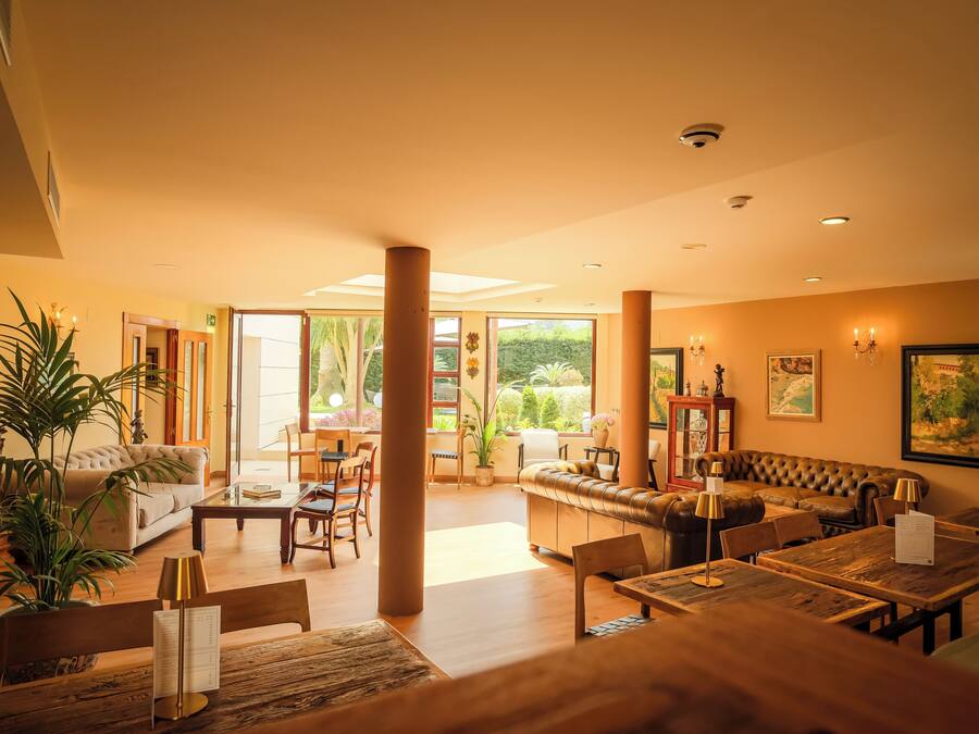 Un hall de hotel espacioso con sofás, mesas de comedor y sillas, ventanas amplias que dan a zonas verdes, y una combinación de decoración moderna y clásica.
