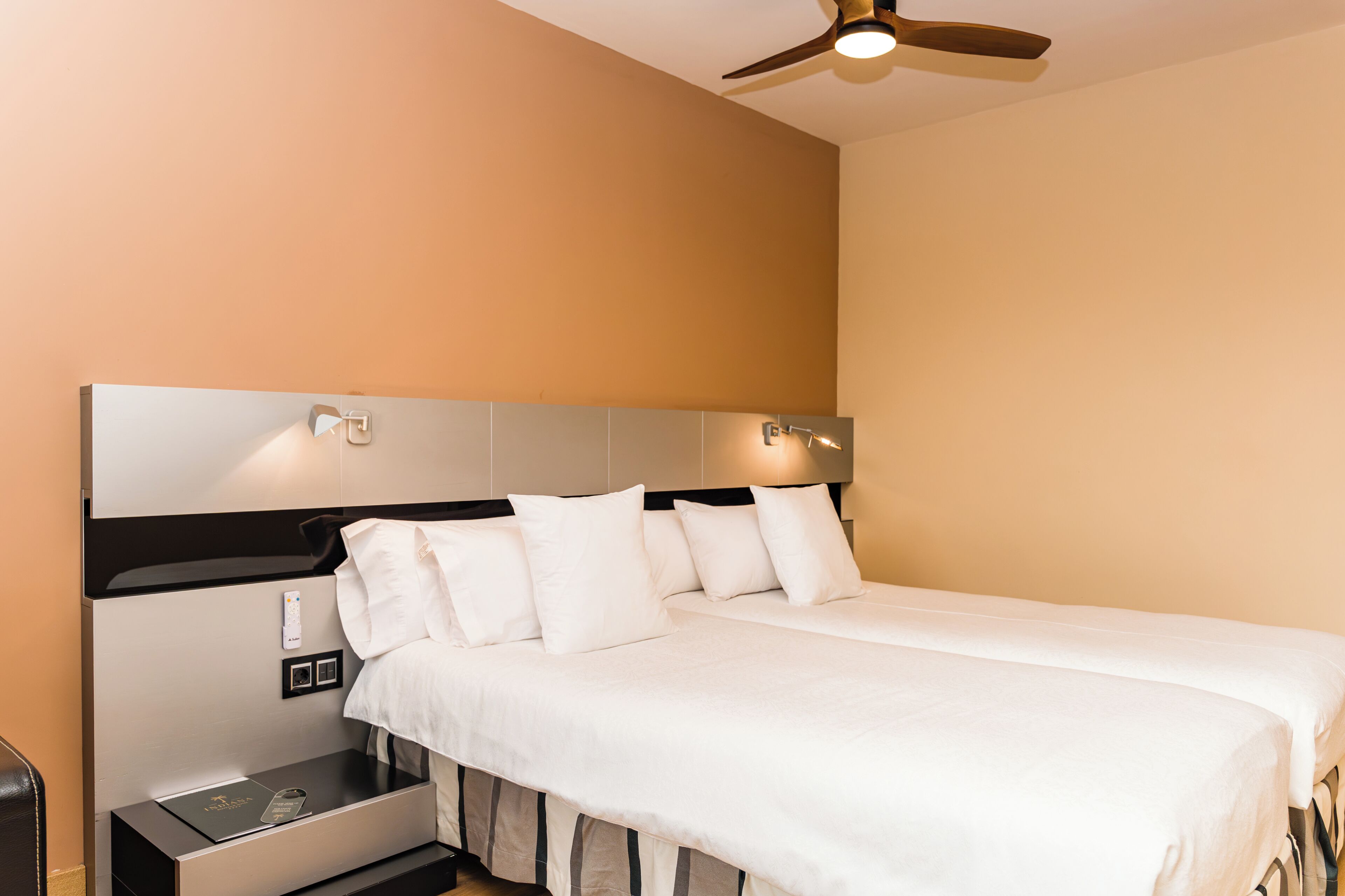 Habitación de hotel con cama, mesita de noche, ventilador de techo y lámpara fijada a la pared.