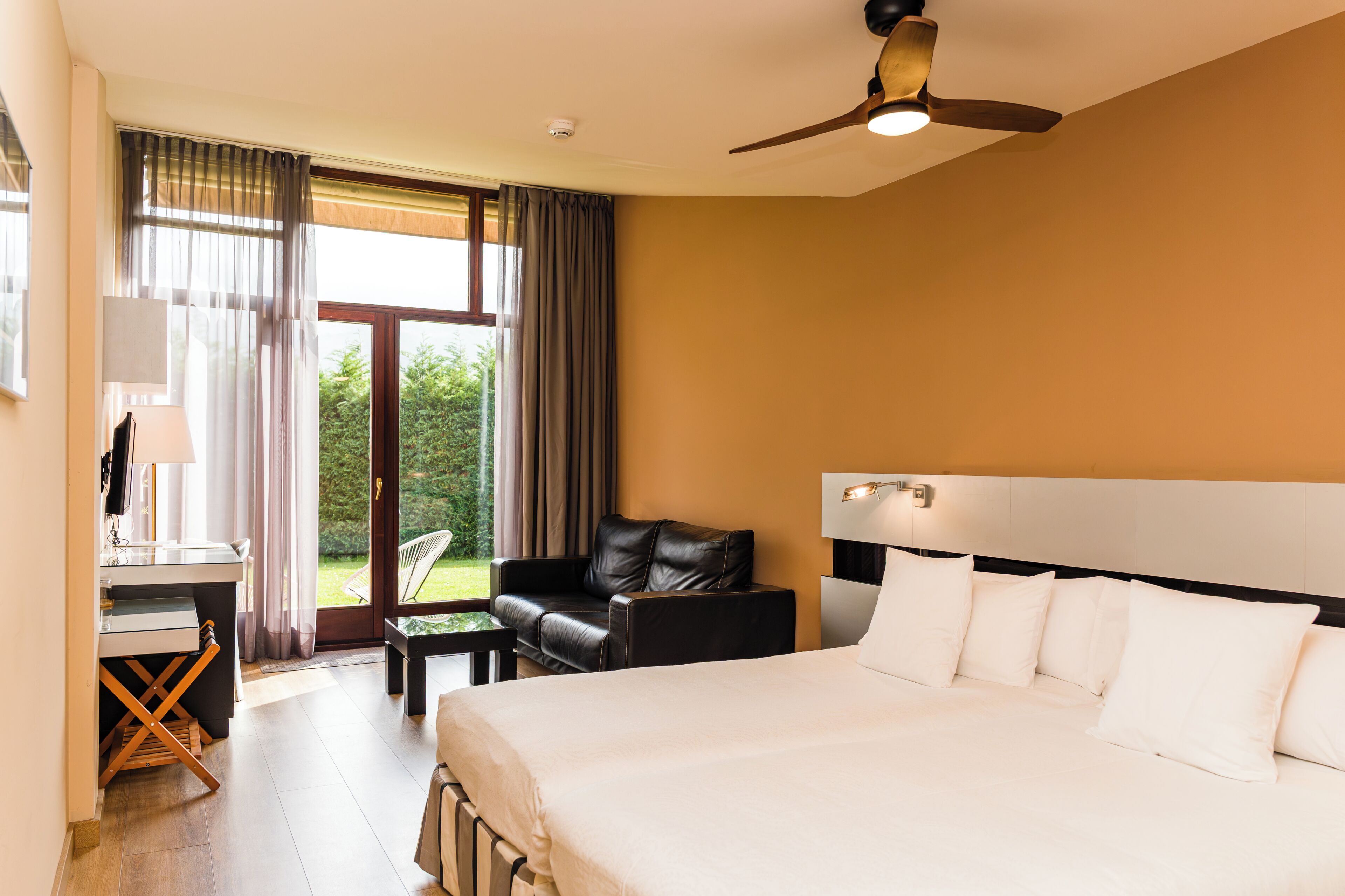 A hotel room with a bed, a desk, a chair, a ceiling fan, and a view of the outdoors.