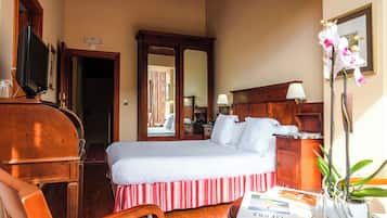 Habitación de hotel con cama, una silla de madera, un escritorio con lámpara, televisor y una orquídea en maceta.