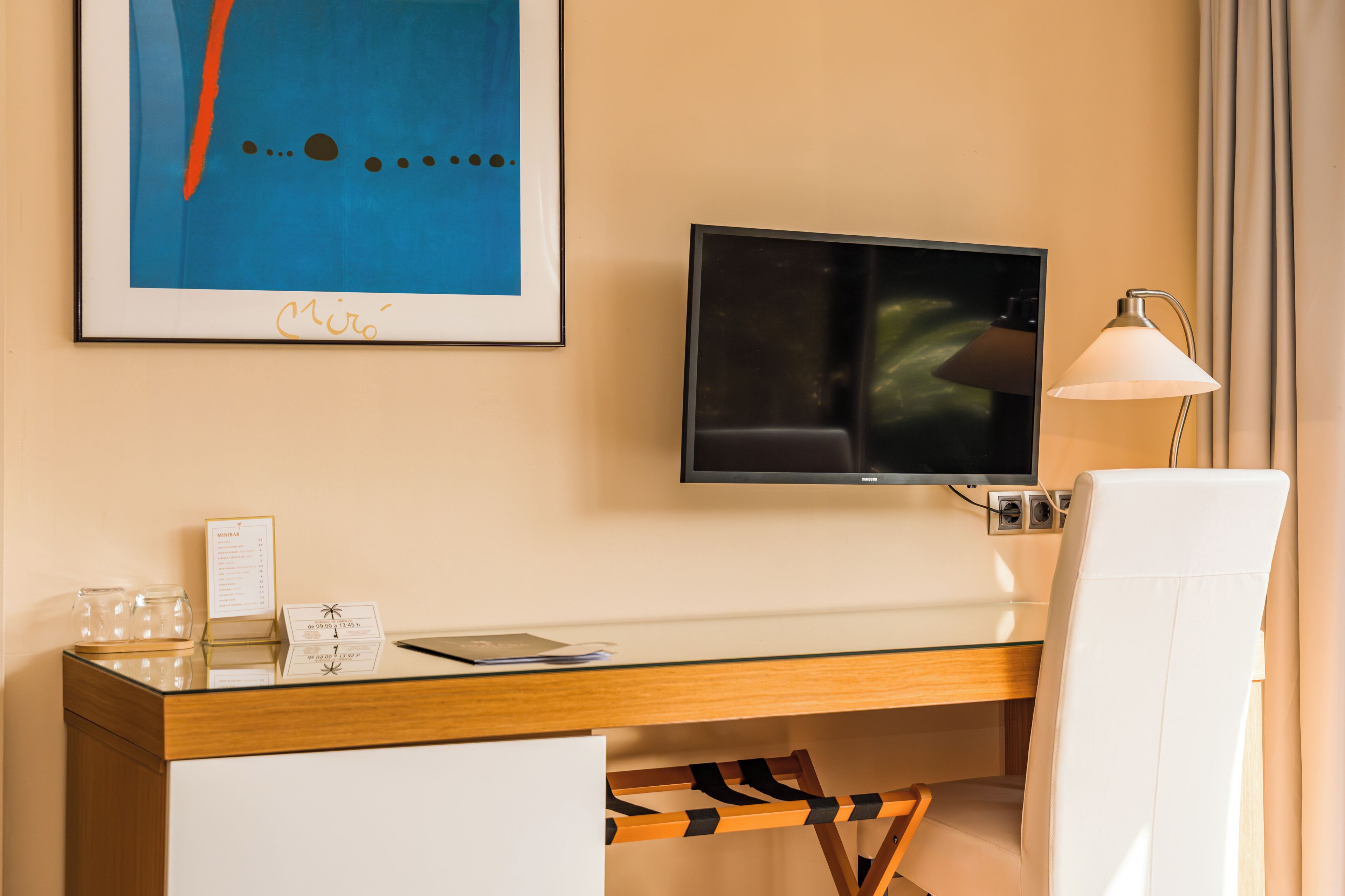 Habitación de hotel con escritorio, televisor montado en la pared, lámpara y una obra de arte enmarcada sobre el escritorio.