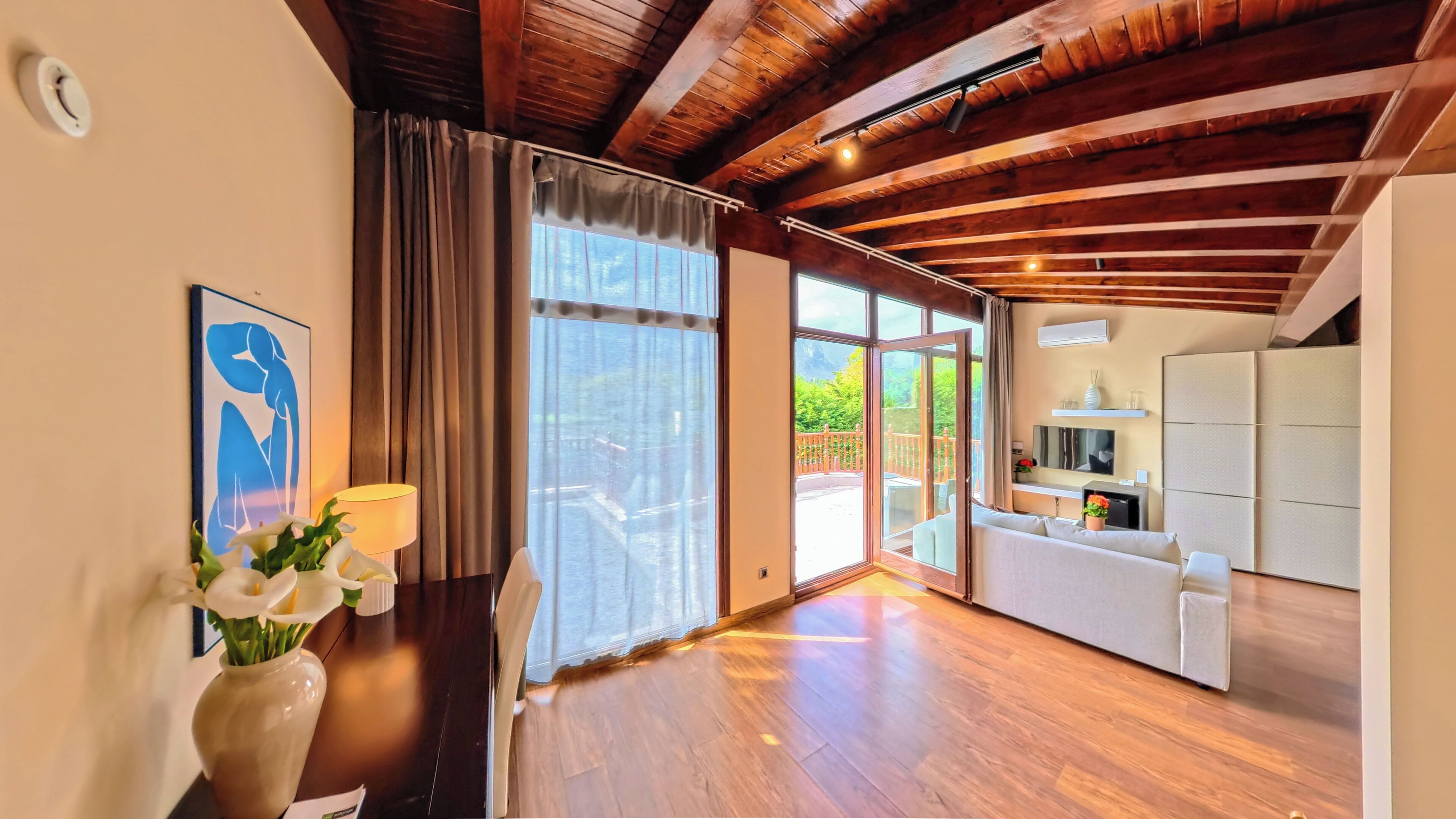 Una habitación amplia con suelo de madera, un ventanal grande, un sofá, una mesa de comedor y un armario blanco.
