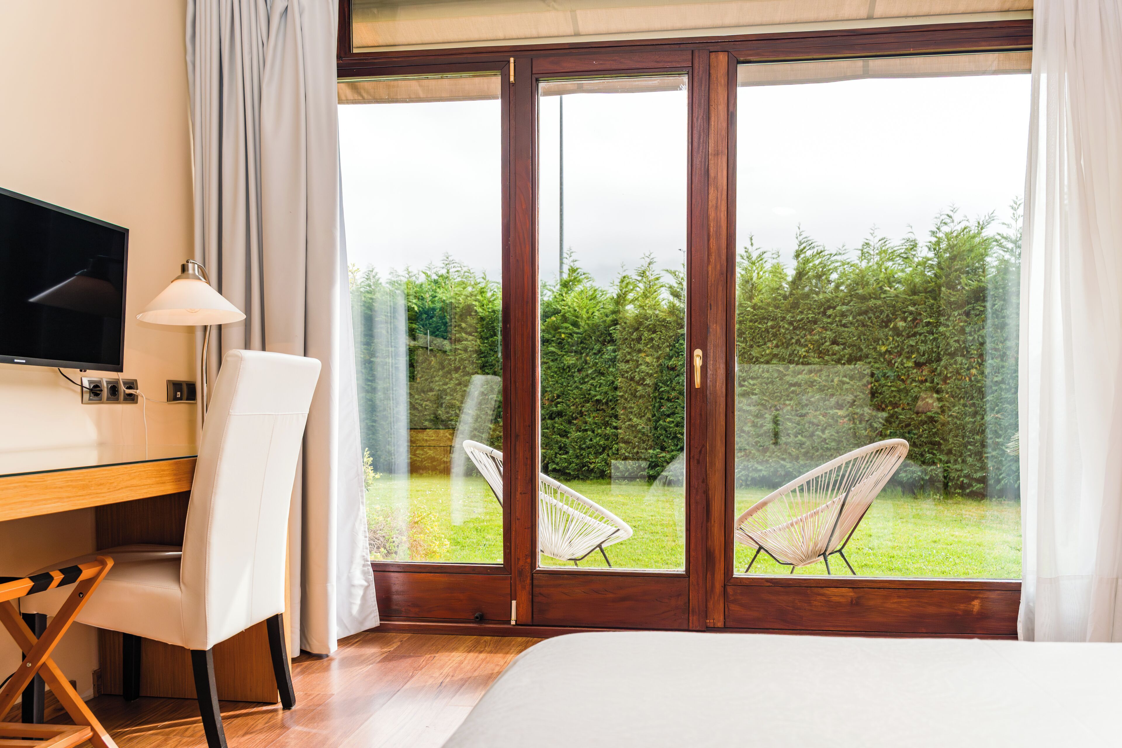 A bedroom with a large sliding glass door leading to a garden. There is a bed, a desk with a chair, and a TV mounted on the wall.