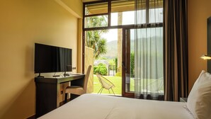 A hotel room with a bed, a desk with a television, a chair, and a view of a garden and mountains through a large window. - Indiana Boutique Hotel (Llanes)