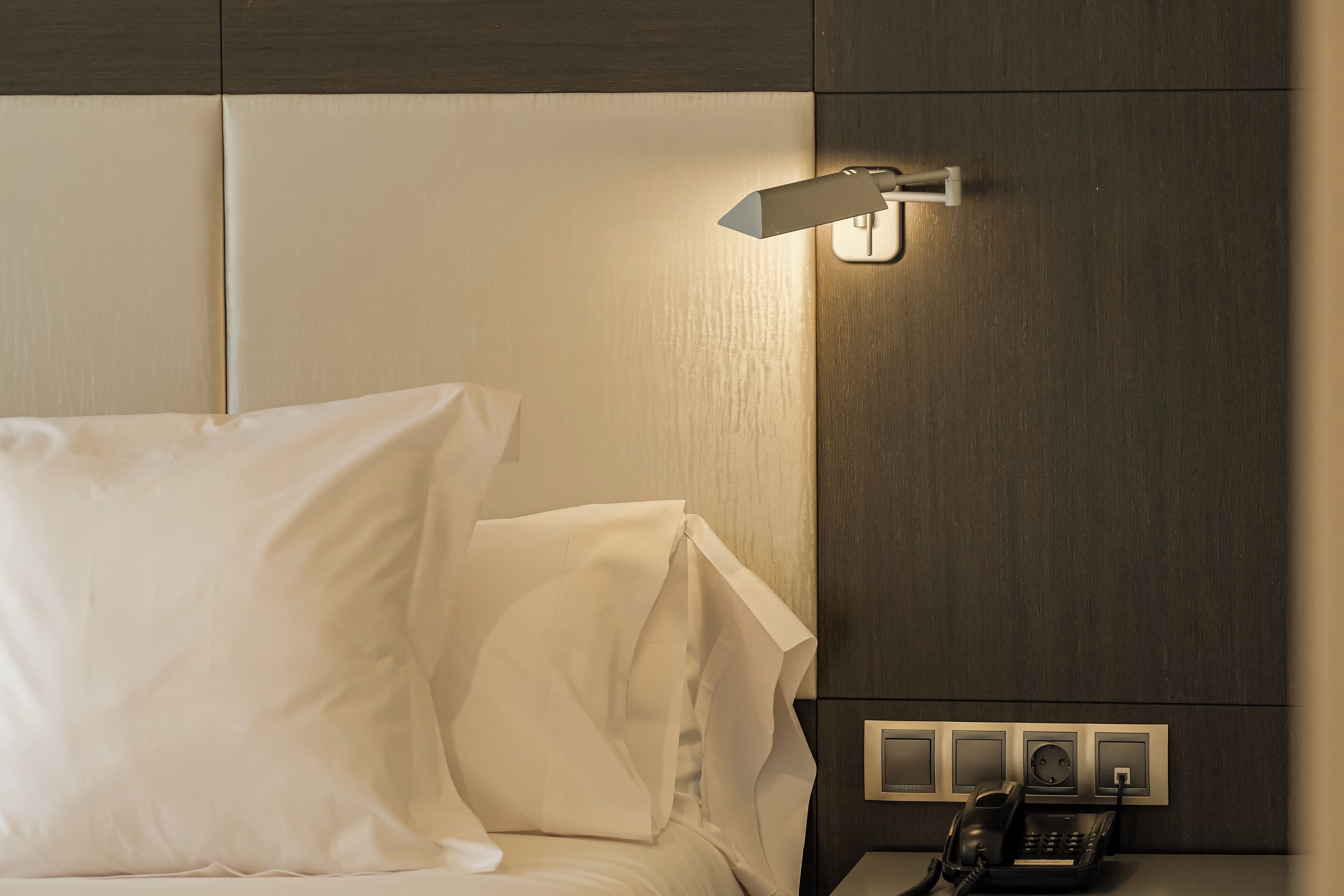 A hotel room with a bed, a bedside lamp, a telephone, and a wall socket.