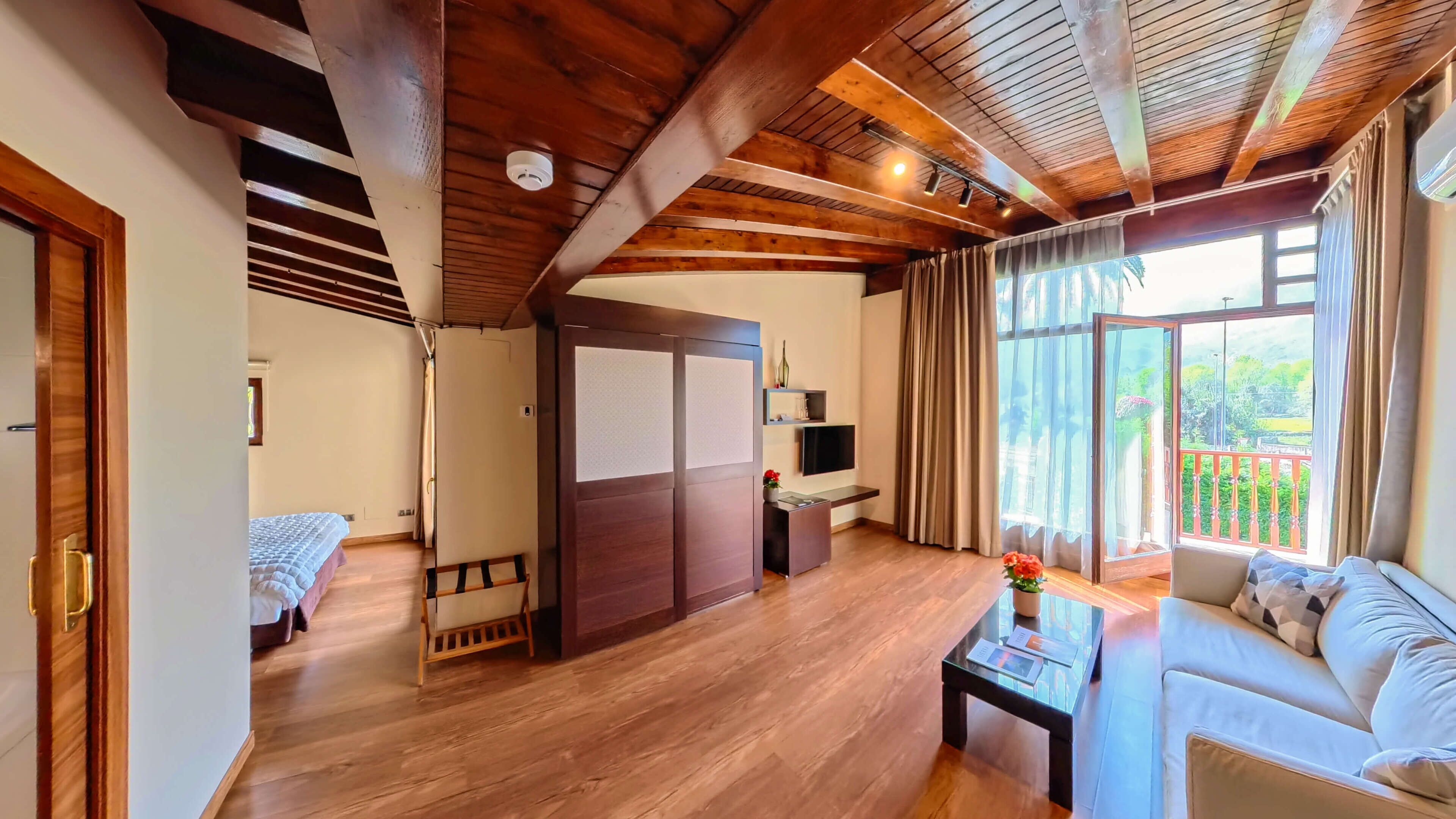 A spacious room with a wooden ceiling, a sofa, a TV, and a balcony with curtains.