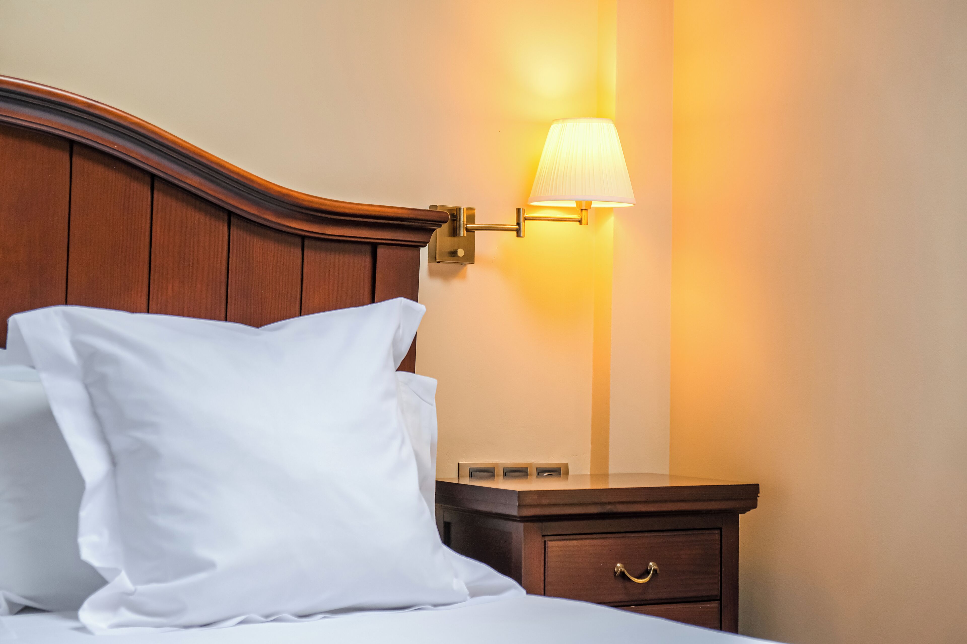 A neatly made bed with white linens and a wooden headboard, a bedside table with a lamp, and a small drawer.