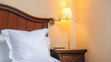 A neatly made bed with white linens and a wooden headboard, a bedside table with a lamp, and a small drawer.