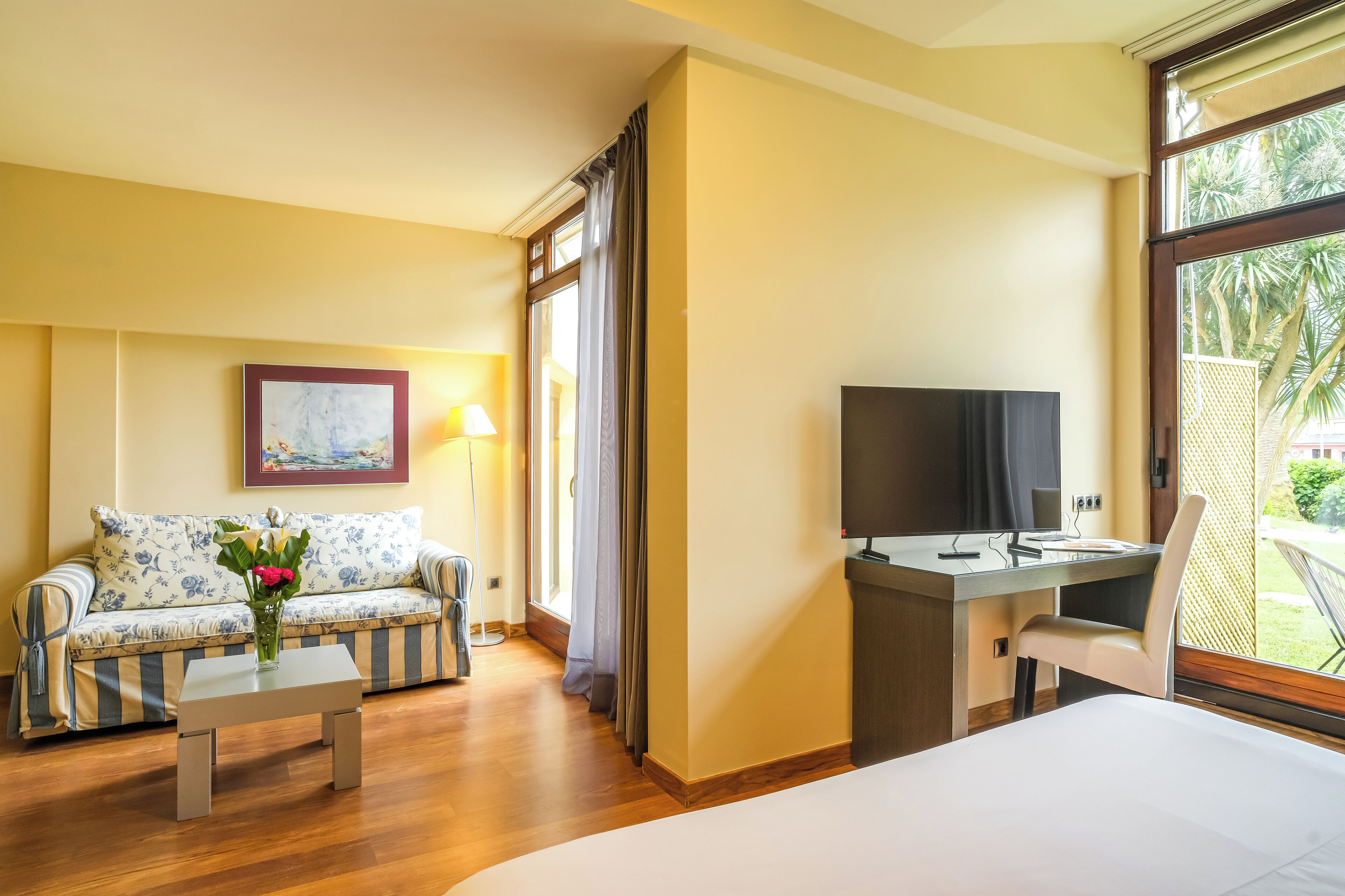 A hotel room with a bed, a sofa, a desk, a TV, and a window with a view of greenery.