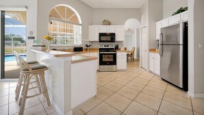 A modern kitchen with white cabinetry, a stainless steel refrigerator, a microwave, and an oven. There is a bar area with stools and a view of the outdoors. - ⛱  Lake View - 4 Bed - Sunridge Wood - South Facing Pool Home ✈ (Davenport)