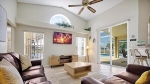 A spacious living room with a brown leather sofa, a wooden coffee table, and a large TV screen. There is a ceiling fan, a window with a view of the outdoors, and a door leading to a balcony. - ⛱  Lake View - 4 Bed - Sunridge Wood - South Facing Pool Home ✈ (Davenport)