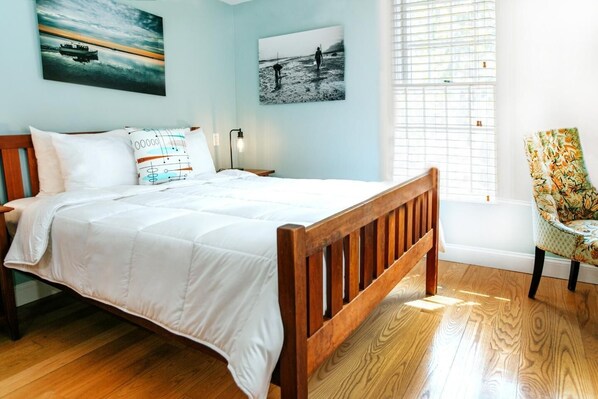 A bedroom with a wooden bed, white bedding, a chair with a floral pattern, and two wall-mounted pictures.