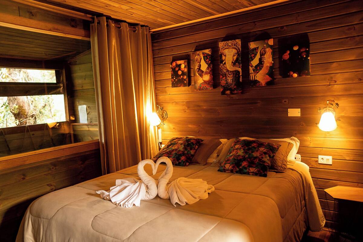 A wooden cabin room with a bed, two wall-mounted lamps, and a window with curtains.