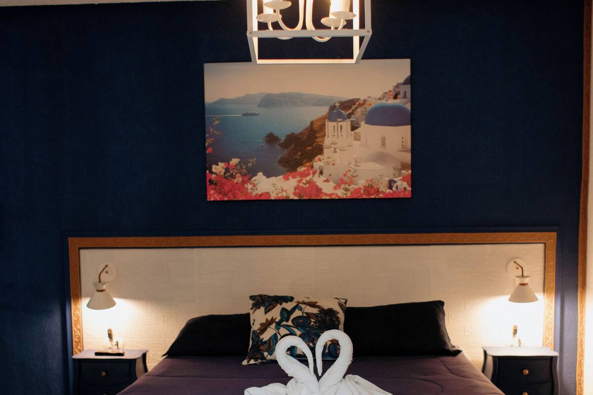A bedroom with a large bed, two bedside tables with lamps, a headboard, a wall-mounted picture, and a decorative swan towel centerpiece.