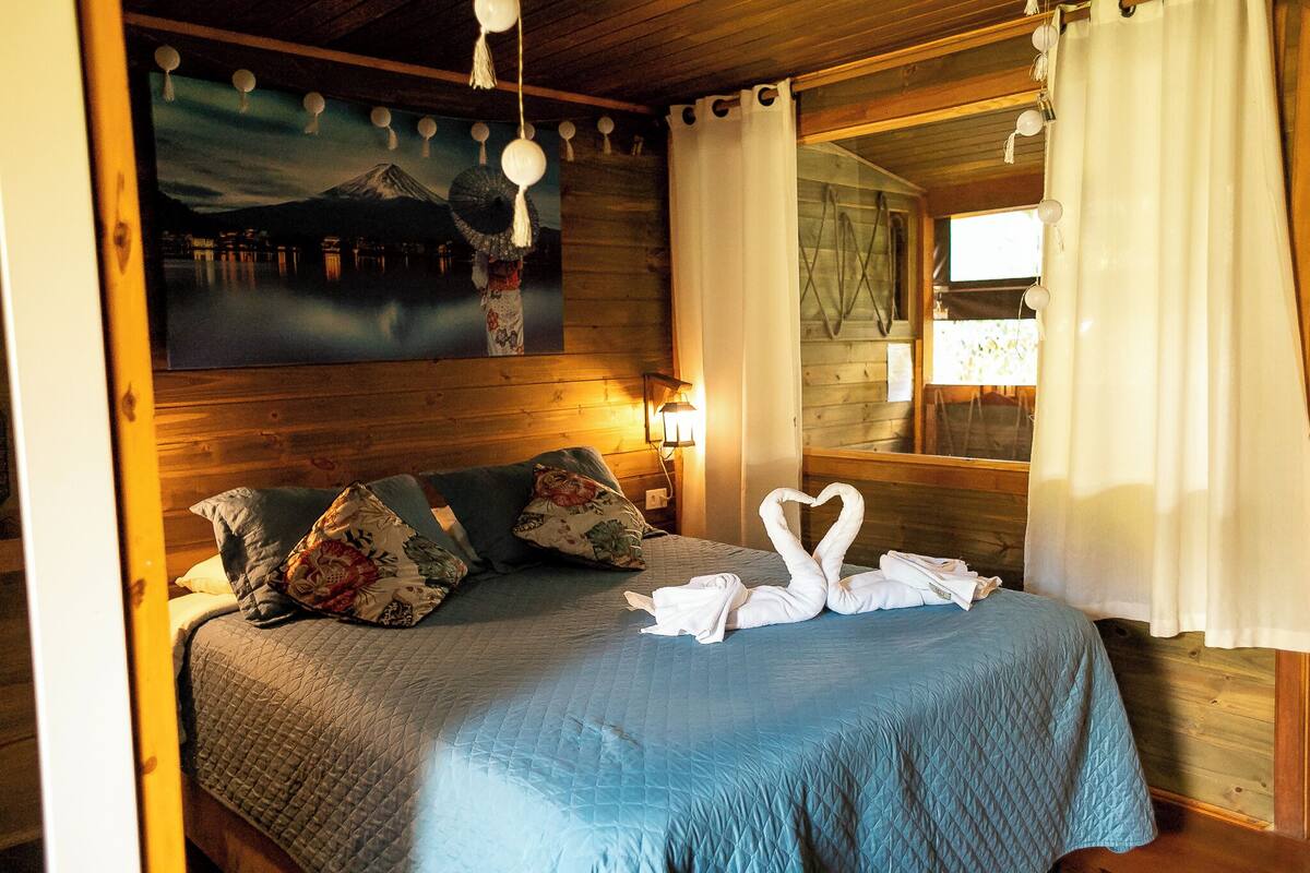 A wooden cabin room with a bed, pillows, a painting of a mountain, and a window with curtains.