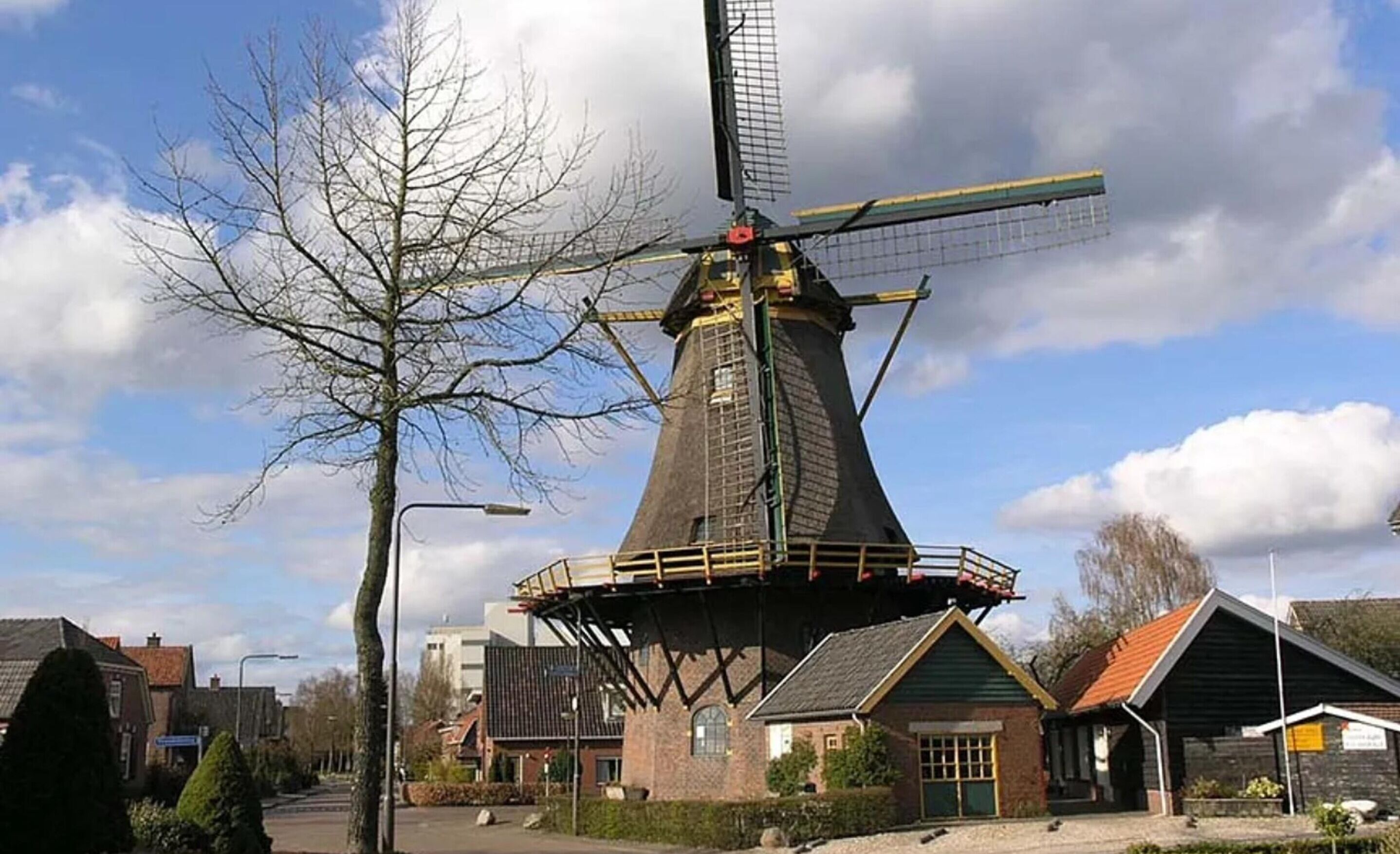 A traditional windmill with a conical roof and a large sail, surrounded by residential buildings and a leafless tree.