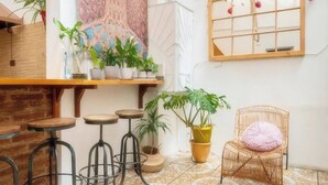 A cozy interior with a high ceiling, a hanging light fixture, a large tapestry, a wooden-framed window, and a wicker chair with a cushion. - Hostal Casa Fibonacci (Valparaíso)