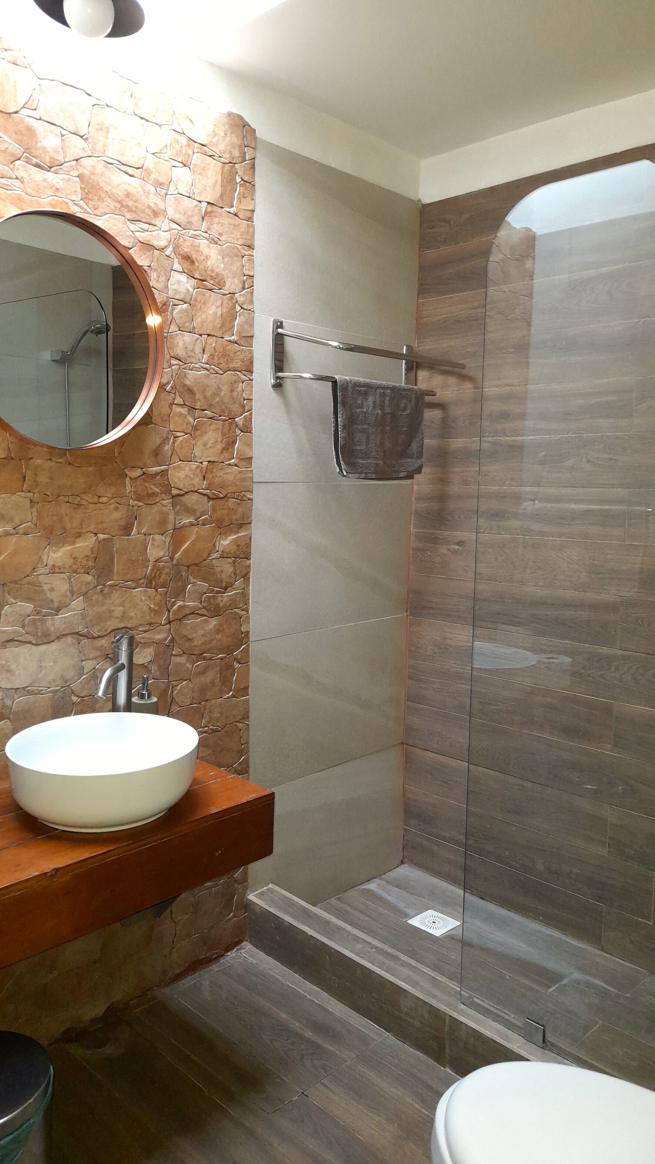 Un baño con pared de piedra, un espejo redondo, un lavabo, un perchero para toallas y una zona de ducha.