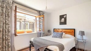 A bedroom with a wooden bed, a window with curtains, a bedside table with a lamp, and a wall-mounted picture. - Hostal Casa Fibonacci (Valparaíso)