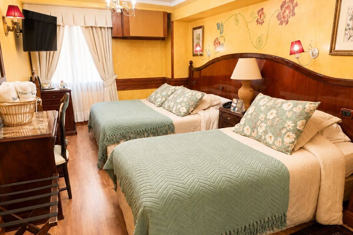 A hotel room with two beds, a desk, a chair, a chandelier, and a patterned rug.