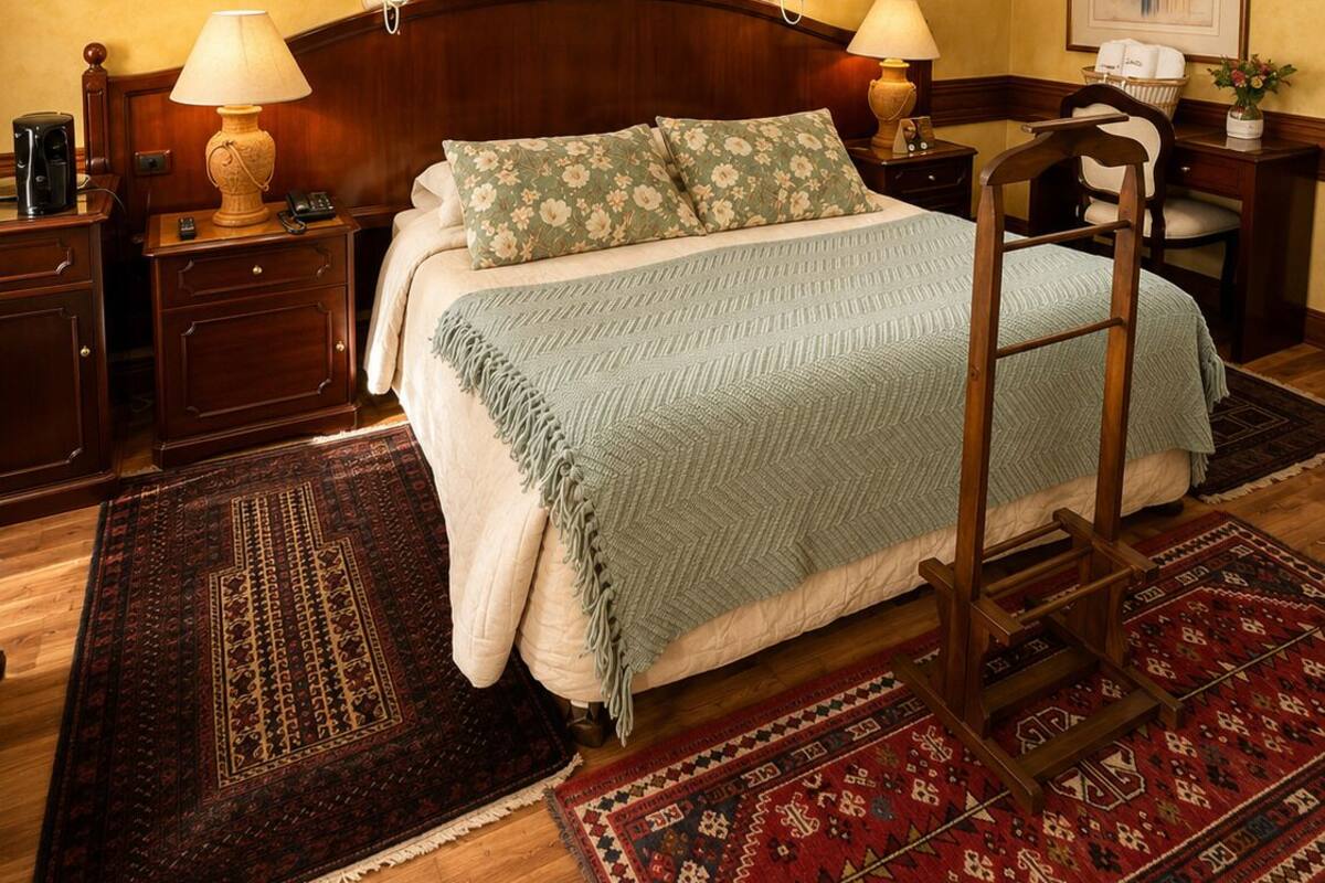 A hotel room with a wooden headboard, a bed with floral bedding, two bedside tables with lamps, a chandelier, and a large fruit painting on the wall.