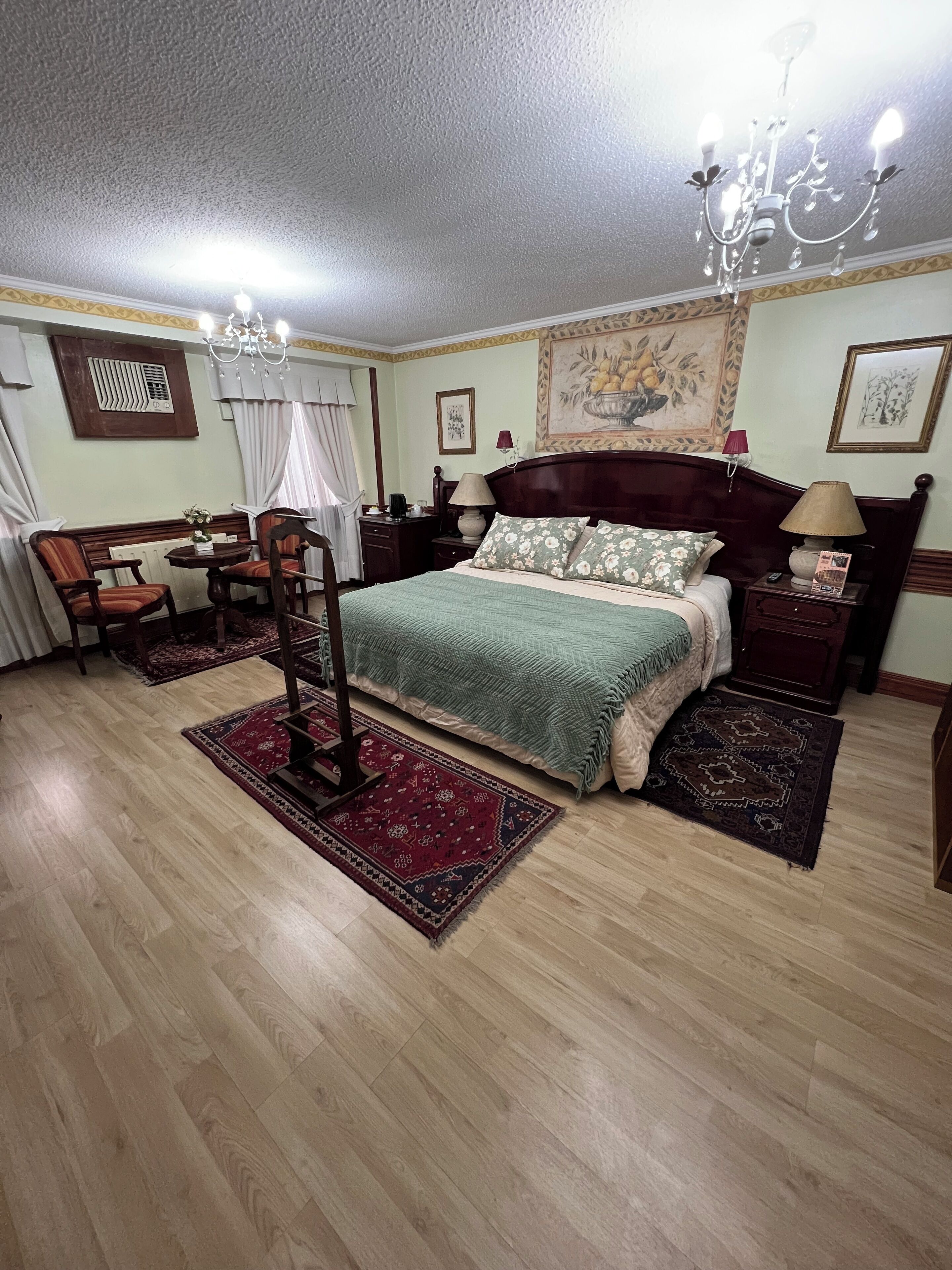 Un dormitorio con cama, candelabro, mesa con sillas, radiador y un cuadro en la pared.