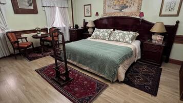 Un dormitorio con cama, candelabro, mesa con sillas, radiador y un cuadro en la pared.