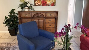 A living room with a blue armchair, a wooden cabinet with decorative handles, a large floral painting, and potted plants. - Peaceful & Pet Friendly (Gainesville)