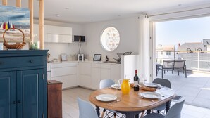 Une cuisine moderne avec des meubles blancs, une table à manger dressée pour deux personnes et une vue sur la ville par une grande fenêtre.