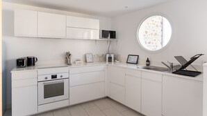 Une cuisine moderne avec des meubles blancs, un four encastré, un four à micro-ondes et un évier. Il y a une fenêtre offrant une vue agréable, ainsi qu'une petite étagère où sont rangés des ustensiles de cuisine.