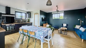 A modern kitchen with dark cabinetry, a dining area with a table and chairs, and a living room with a blue sofa and a TV.