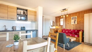 A modern kitchen with wooden cabinets, a dining table with chairs, a red sofa, and a small table with a lamp.