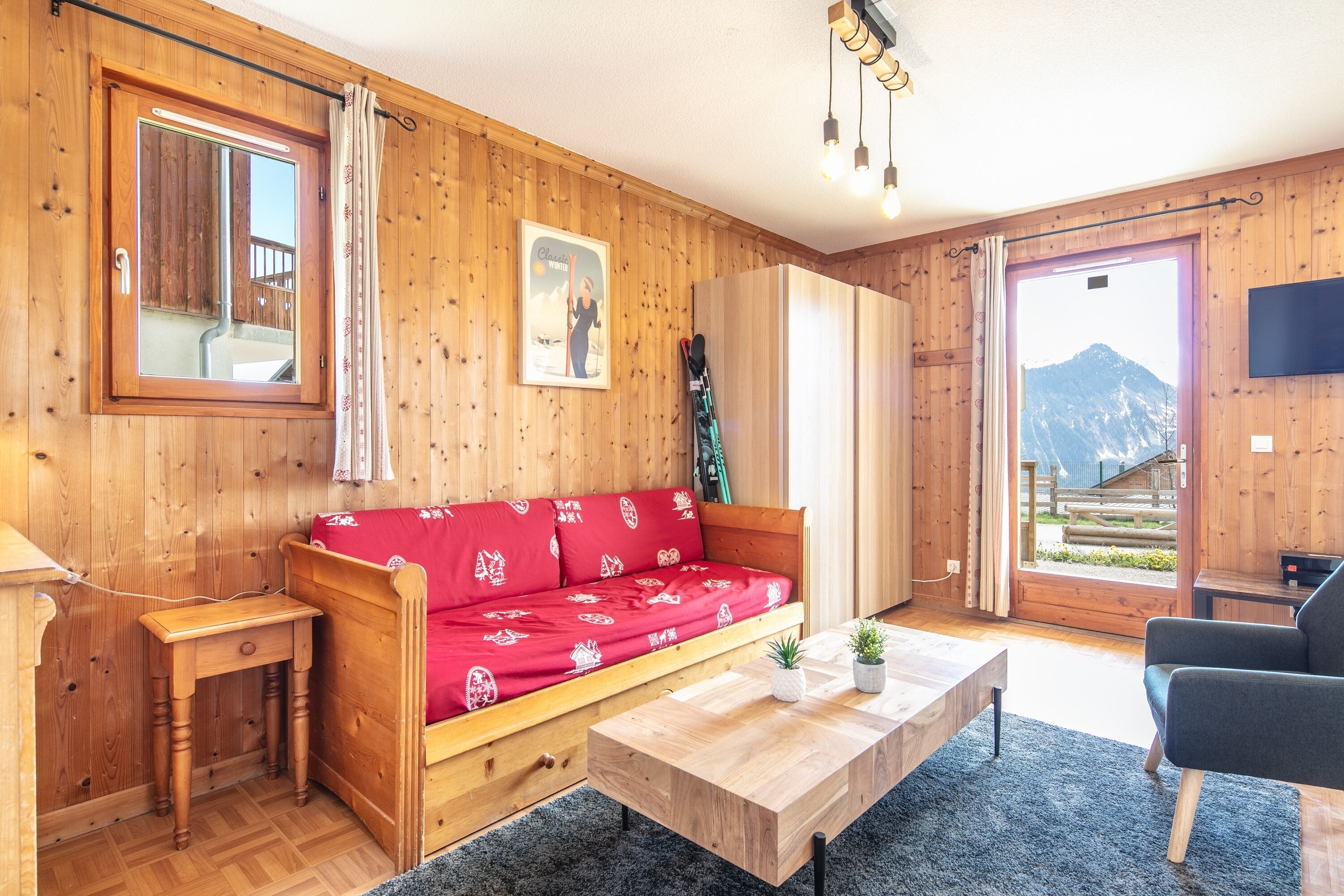 A wooden-furnished room with a red sofa, a wooden table, a TV, and a mountain view.