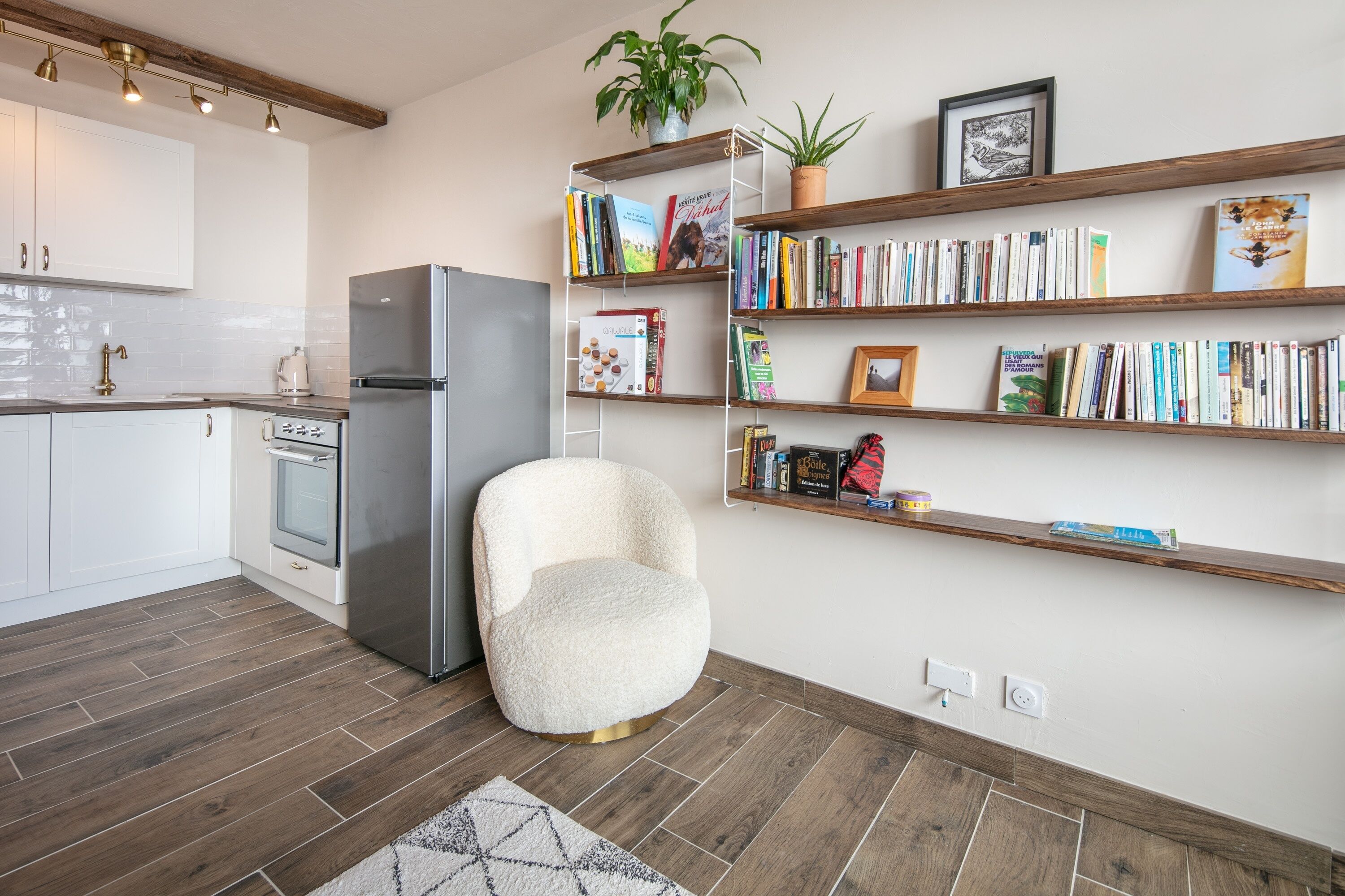 A modern kitchen with white cabinets, a stainless steel refrigerator, and a white chair. There are wooden shelves with books and a potted plant.
